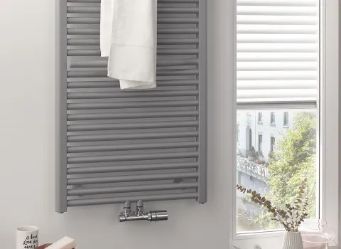 In einem hellen Badezimmer hängt ein grauer HSK Line Plus Heizkörper mit einem weißen Handtuch daran. 