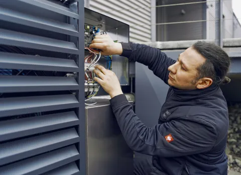Ein Techniker überprüft eine Elektroinstallation an einer Außeneinheit, während er sorgfältig Kabel in einem offenen Schaltkasten anschließt.