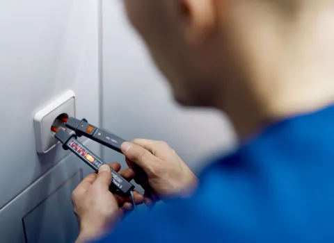 Ein Elektriker SHK in einem blauen Arbeitshemd überprüft mit einem Spannungsprüfer eine Steckdose.