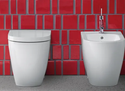 Ein elegantes weißes WC und Bidet, minimalistisch gestaltet, vor einer Wand aus roten Kacheln in kräftigem Glanz.