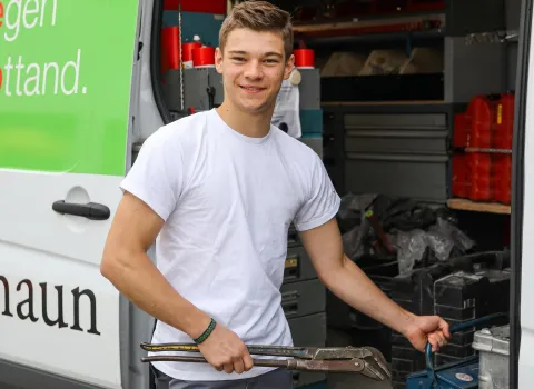 Der lächelnde Alexander Hierl, ein junger Mann in weißem T-Shirt, hält eine große Rohrzange und steht vor dem offenen Laderaum eines weißen Lieferwagens. Im Inneren des Wagens sind Werkzeuge und rote Behälter auf Regalen zu erkennen. Am linken Bildrand ist ein grünes Schild mit weißer Schrift am Fahrzeug zu sehen.