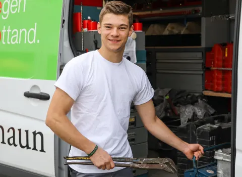 Der lächelnde Alexander Hierl, ein junger Mann in weißem T-Shirt, hält eine große Rohrzange und steht vor dem offenen Laderaum eines weißen Lieferwagens. Im Inneren des Wagens sind Werkzeuge und rote Behälter auf Regalen zu erkennen. Am linken Bildrand ist ein grünes Schild mit weißer Schrift am Fahrzeug zu sehen.
