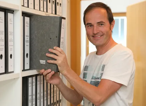Andreas Simon lächelt freundlich, während er in einem Büro oder Archiv einen dunkelgrauen Ordner aus einem weißen Regal zieht, das mit weiteren Aktenordnern gefüllt ist.