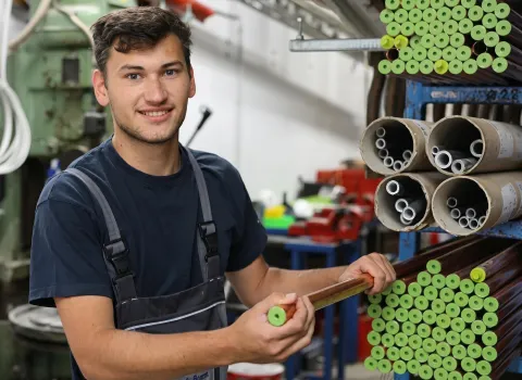 Ein lächelnder junger Mann, Bastian Nutz, in dunkelblauer Arbeitskleidung, hält ein kupferfarbenes Rohr mit hellgrüner Schutzkappe in den Händen. Er steht in einer Lagerhalle, umgeben von Regalen voller ähnlicher Rohre mit grünen Kappen und Kartonrollen mit kleineren Metallrohren.