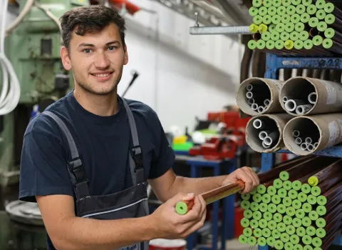 Ein lächelnder junger Mann, Bastian Nutz, in dunkelblauer Arbeitskleidung, hält ein kupferfarbenes Rohr mit hellgrüner Schutzkappe in den Händen. Er steht in einer Lagerhalle, umgeben von Regalen voller ähnlicher Rohre mit grünen Kappen und Kartonrollen mit kleineren Metallrohren.