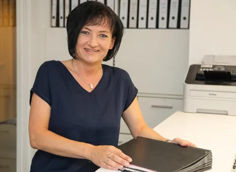 Brigitte Bschor sitzt an einem weißen Schreibtisch, lächelt freundlich in die Kamera und hält einen schwarzen Ordner. Sie trägt ein dunkelblaues Oberteil und eine silberne Halskette. Im Hintergrund sind Aktenordner und ein Drucker zu sehen.
