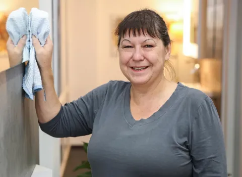 Christine Forster lächelt direkt in die Kamera, während sie mit einem hellblauen, waffelgemusterten Reinigungstuch eine spiegelnde Oberfläche abwischt. Sie trägt ein graues V-Ausschnitt-T-Shirt.