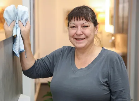 Christine Forster lächelt direkt in die Kamera, während sie mit einem hellblauen, waffelgemusterten Reinigungstuch eine spiegelnde Oberfläche abwischt. Sie trägt ein graues V-Ausschnitt-T-Shirt.