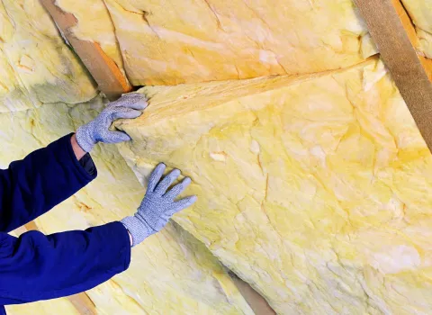 Ein Handwerker mit Handschuhen schiebt gelbe Mineralwolle zur Dämmung des Gebäudes in die Hauswand hinein.