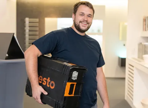 Daniel Schertel lächelt freundlich in die Kamera, während er einen schwarzen Testo-Koffer mit orangefarbenen Akzenten unter dem rechten Arm trägt. Er trägt ein dunkelblaues T-Shirt und hat einen kurzen Bart. Im Hintergrund ist ein moderner Innenraum mit hellen Wänden zu sehen.