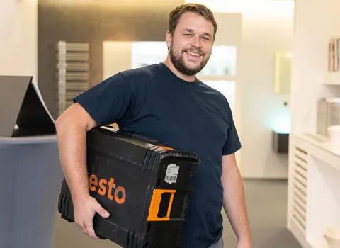 Daniel Schertel lächelt freundlich in die Kamera, während er einen schwarzen Testo-Koffer mit orangefarbenen Akzenten unter dem rechten Arm trägt. Er trägt ein dunkelblaues T-Shirt und hat einen kurzen Bart. Im Hintergrund ist ein moderner Innenraum mit hellen Wänden zu sehen.