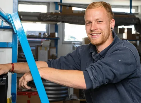 Dominic Stüwe, ein lächelnder Mann mit rötlich-blondem Haar und Bart, trägt ein dunkelblaues Poloshirt und lehnt sich an eine blaue Metallstruktur in einem Lagerraum. Er hat einen Ohr-Tunnel im linken Ohr und ein rotes Armband am rechten Handgelenk.