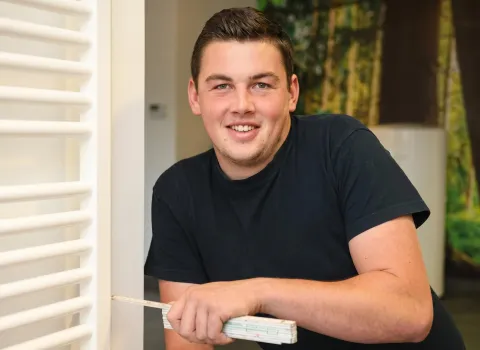 Florian Sußner, ein junger Mann mit kurzem, dunklem Haar und einem Lächeln, trägt ein schwarzes T-Shirt und hält einen gefalteten Gliedermaßstab in seiner rechten Hand. Er lehnt sich an einen weißen Lamellenheizkörper. Der Hintergrund ist unscharf mit grünen und braunen Farbakzenten.
