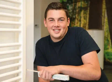 Florian Sußner, ein junger Mann mit kurzem, dunklem Haar und einem Lächeln, trägt ein schwarzes T-Shirt und hält einen gefalteten Gliedermaßstab in seiner rechten Hand. Er lehnt sich an einen weißen Lamellenheizkörper. Der Hintergrund ist unscharf mit grünen und braunen Farbakzenten.