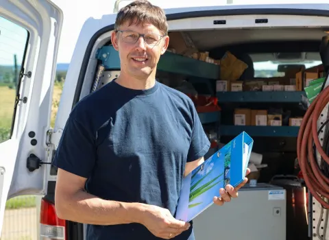 Franz Schöll, ein Mann mit Brille und dunkelblauem T-Shirt, lächelt und hält einen blauen Ordner. Er steht vor dem geöffneten Heck eines weißen Transporters mit Regalen voller Kartons und Werkzeugen.