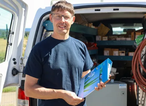 Franz Schöll, ein Mann mit Brille und dunkelblauem T-Shirt, lächelt und hält einen blauen Ordner. Er steht vor dem geöffneten Heck eines weißen Transporters mit Regalen voller Kartons und Werkzeugen.