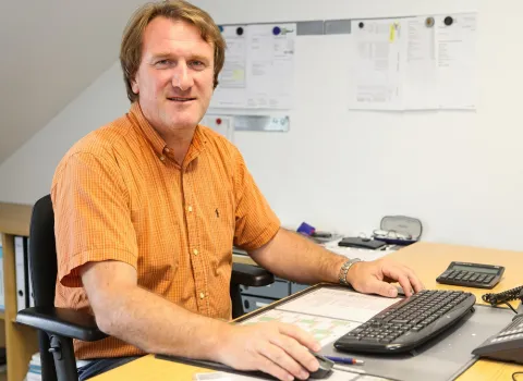 Günther Waffler sitzt an einem hellbraunen Schreibtisch in einem Büro, trägt ein orangefarbenes kariertes Hemd und lächelt in die Kamera. Seine Hände liegen auf einer Maus und Tastatur, während im Hintergrund Büroakten und eine weiße Wand mit Dokumenten zu sehen sind.