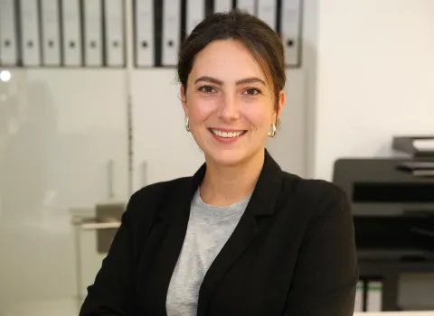 Jennifer Rinke lächelt freundlich in die Kamera. Sie hat dunkle, zurückgebundene Haare, braune Augen und trägt silberne Creolen, ein hellgraues T-Shirt und einen schwarzen Blazer. Im unscharfen Hintergrund sind Aktenordner und eine Glastrennwand in einem Büro zu sehen.