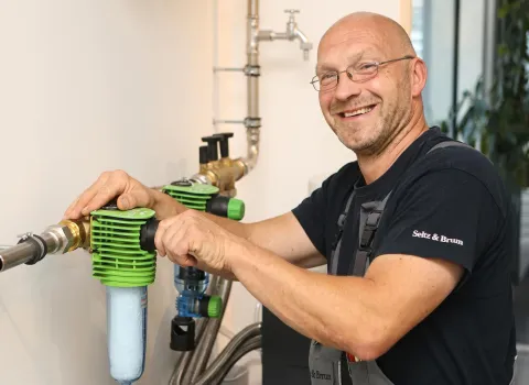 Jens Kreutzer, ein lächelnder Mann mit Brille, hält eine grüne Wasserfilteranlage mit einem blauen Filter in den Händen. Er trägt ein schwarzes T-Shirt und graue Latzhosen vor einer Wand mit Wasserleitungen.