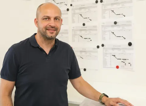 Jochen Schreiber, ein lächelnder Mann mit kurzen Haaren und Bart, trägt ein dunkelblaues Poloshirt. Er steht vor einer weißen Wand, an der mit schwarzen und einem roten Magneten mehrere Zeitpläne oder Diagramme befestigt sind. Seine rechte Hand ruht auf einem weißen Tisch, während er am linken Handgelenk eine grüne Digitaluhr trägt.