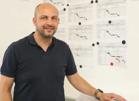 Jochen Schreiber, ein lächelnder Mann mit kurzen Haaren und Bart, trägt ein dunkelblaues Poloshirt. Er steht vor einer weißen Wand, an der mit schwarzen und einem roten Magneten mehrere Zeitpläne oder Diagramme befestigt sind. Seine rechte Hand ruht auf einem weißen Tisch, während er am linken Handgelenk eine grüne Digitaluhr trägt.