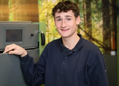 Der junge Mann Johannes Heider mit lockigem, braunem Haar und einem Lächeln blickt direkt in die Kamera. Er trägt ein dunkelblaues Poloshirt und hat seine rechte Hand auf einem grauen Gerät mit einem dunklen Bildschirm platziert. Im Hintergrund sind unscharfe grüne und braune Formen zu erkennen, die an Bäume oder Blätter erinnern.
