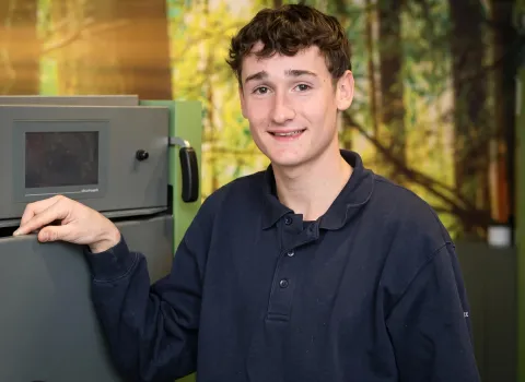 Der junge Mann Johannes Heider mit lockigem, braunem Haar und einem Lächeln blickt direkt in die Kamera. Er trägt ein dunkelblaues Poloshirt und hat seine rechte Hand auf einem grauen Gerät mit einem dunklen Bildschirm platziert. Im Hintergrund sind unscharfe grüne und braune Formen zu erkennen, die an Bäume oder Blätter erinnern.