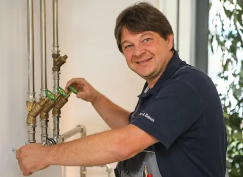 Der lächelnde Heizungsinstallateur Jürgen Rössel in blauer Polohemd und grauer Latzhose dreht ein grünes Ventil an einem System aus verchromten Rohren und Messingventilen, das an einer weißen Wand montiert ist.