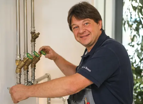 Der lächelnde Heizungsinstallateur Jürgen Rössel in blauer Polohemd und grauer Latzhose dreht ein grünes Ventil an einem System aus verchromten Rohren und Messingventilen, das an einer weißen Wand montiert ist.