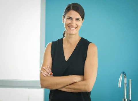 Julia Haller lächelt direkt in die Kamera, ihre Arme sind verschränkt. Sie trägt ein schwarzes ärmelloses Oberteil, Perlohrringe und einen auffälligen silbernen Ring mit rotem Stein an ihrer linken Hand. Im Hintergrund ist eine türkise Wand und ein weißes Fenster zu sehen.