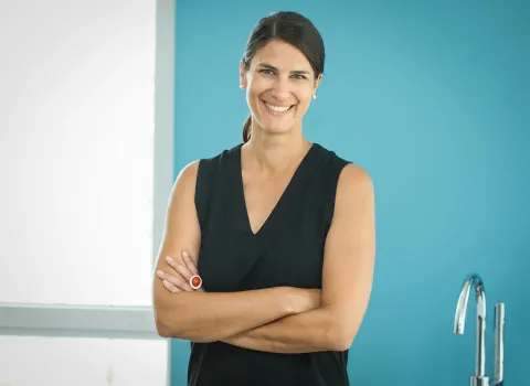 Julia Haller lächelt direkt in die Kamera, ihre Arme sind verschränkt. Sie trägt ein schwarzes ärmelloses Oberteil, Perlohrringe und einen auffälligen silbernen Ring mit rotem Stein an ihrer linken Hand. Im Hintergrund ist eine türkise Wand und ein weißes Fenster zu sehen.