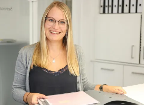 Katja Foistner lächelt in die Kamera, während sie in einem Büro einen grauen Aktenordner mit rosa Dokumenten hält. Sie trägt eine graue Strickjacke, ein schwarzes Top mit Spitze und eine silberne Halskette mit rundem Anhänger. Im Hintergrund sind weiße Büroschränke und ein beschrifteter Glastürbereich sichtbar.