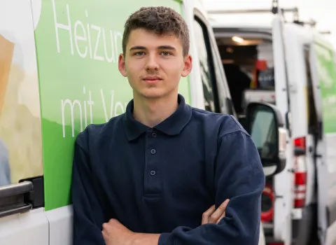Louis Distler, ein junger Mann mit brauen Haaren und einem dunkelblauen Poloshirt, lehnt mit verschränkten Armen an der Seite eines weißen Lieferwagens. Ein grüner Bereich auf dem Lieferwagen ist mit weißer Schrift beschriftet. Im Hintergrund sind die geöffneten hinteren Türen des Transporters mit sichtbarem Inhalt zu sehen.