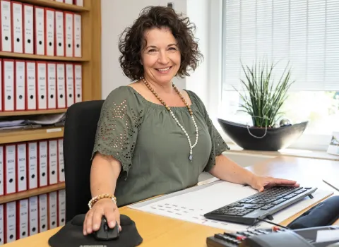 Manuela Bittner sitzt lächelnd an einem hellbraunen Schreibtisch in ihrem Büro, die rechte Hand auf einer Computermaus und die linke Hand auf einer Tastatur. Sie trägt ein grünes Oberteil mit Lochmusterärmeln und zwei Halsketten, eine aus Holzperlen und eine aus weißen Perlen mit einem blauen Anhänger. Im Hintergrund sind ein Regal mit roten Aktenordnern und ein Fenster mit Jalousien und einer Zimmerpflanze zu sehen.