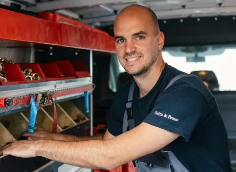Michael Sittel, ein lächelnder Techniker mit Bart und Glatze, trägt ein dunkelblaues Arbeitsshirt mit dem Aufdruck 'Seitz & Braun' und eine graue Latzhose. Er steht in einem Servicefahrzeug, das mit roten Lagerboxen voller Messingfittinge ausgestattet ist, und schaut direkt in die Kamera.