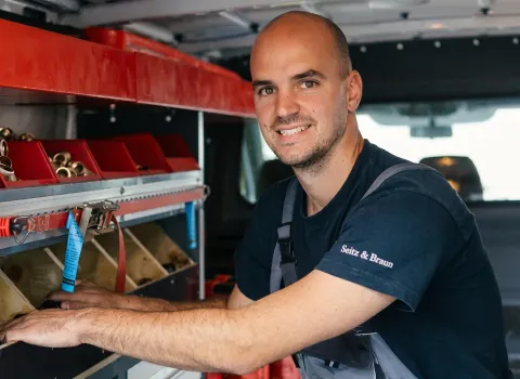 Michael Sittel, ein lächelnder Techniker mit Bart und Glatze, trägt ein dunkelblaues Arbeitsshirt mit dem Aufdruck 'Seitz & Braun' und eine graue Latzhose. Er steht in einem Servicefahrzeug, das mit roten Lagerboxen voller Messingfittinge ausgestattet ist, und schaut direkt in die Kamera.