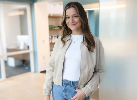Eine lächelnde Michaela Heiselbetz mit langen, hellbraunen Haaren und blonden Strähnen trägt eine beigefarbene Bomberjacke, ein weißes Top und blaue Jeans, während sie mit der linken Hand in der Hosentasche in einem modernen Büroumfeld steht.