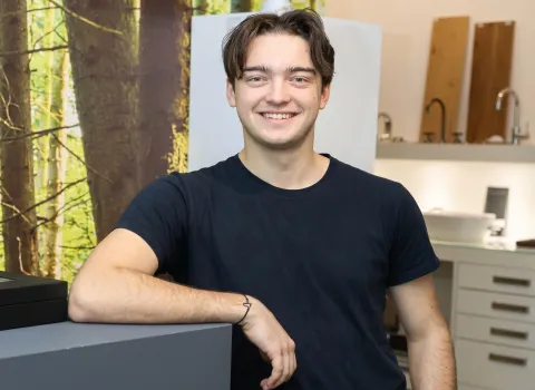 Nico Lell, ein lächelnder junger Mann mit kurzen, dunkelbraunen Haaren, trägt ein dunkelblaues T-Shirt und lehnt mit dem linken Unterarm auf einer dunklen Theke. An seinem linken Handgelenk ist ein dünnes schwarzes Armband zu sehen. Im unscharfen Hintergrund sind Baumstämme und helle Armaturen sowie Holzpaneele erkennbar.