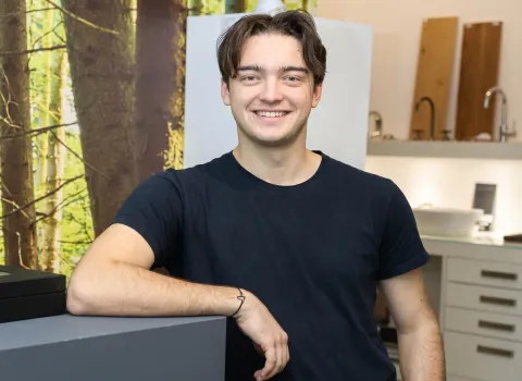 Nico Lell, ein lächelnder junger Mann mit kurzen, dunkelbraunen Haaren, trägt ein dunkelblaues T-Shirt und lehnt mit dem linken Unterarm auf einer dunklen Theke. An seinem linken Handgelenk ist ein dünnes schwarzes Armband zu sehen. Im unscharfen Hintergrund sind Baumstämme und helle Armaturen sowie Holzpaneele erkennbar.