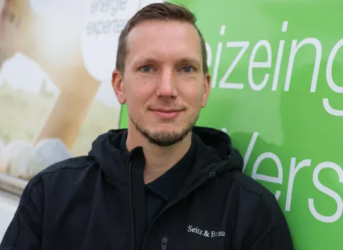 Peter Gruber, ein Mann mit blauen Augen und kurzem Bart, trägt eine schwarze Jacke mit dem weißen Schriftzug "Seitz & Brau" auf der Brust. Er lächelt leicht und blickt direkt in die Kamera. Der Hintergrund ist teilweise hellgrün mit großen weißen Buchstaben und teilweise unscharf beige-weiß mit der Aufschrift "energie experte".