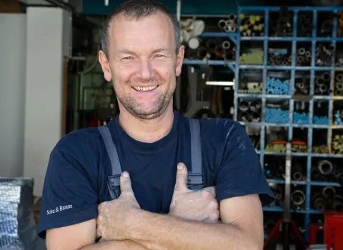 Roland Haas, ein Handwerker, lächelt freundlich in die Kamera, seine Arme sind verschränkt und seine Hände leicht mit hellem Staub bedeckt. Er trägt ein dunkelblaues T-Shirt mit der Aufschrift "Schölz & Braun" auf dem rechten Ärmel und graue Latzhosen. Im Hintergrund sind unscharfe blaue Regale mit verschiedenen Metallrohren und industriellen Materialien zu sehen.