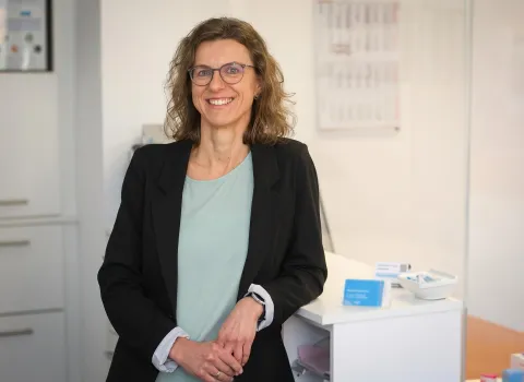 Silke Klebl, eine lächelnde Frau mit brünettem, welligem Haar und Brille, trägt einen schwarzen Blazer und ein mintgrünes Oberteil. Sie steht mit gefalteten Händen an einer weißen Theke in einer hellen, modernen Praxisumgebung.