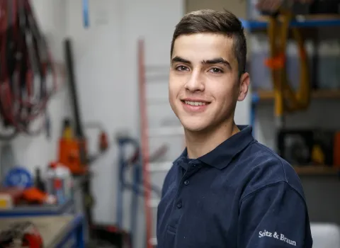 Stefan Freihart, ein lächelnder junger Mann mit kurzen braunen Haaren und dunkelblauem Poloshirt mit dem Schriftzug 'Seltz & Braun', blickt direkt in die Kamera. Im unscharfen Hintergrund sind Werkstattregale und Geräte zu erkennen.