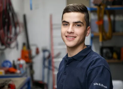 Stefan Freihart, ein lächelnder junger Mann mit kurzen braunen Haaren und dunkelblauem Poloshirt mit dem Schriftzug 'Seltz & Braun', blickt direkt in die Kamera. Im unscharfen Hintergrund sind Werkstattregale und Geräte zu erkennen.