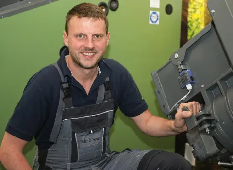 Thomas Blomenhofer, ein lächelnder Mann in dunkelblauem Poloshirt und grau-schwarzer Arbeitslatzhose mit „Solar & Service“-Logo, hockt vor einer grünen Maschine und hält ein graues Maschinenteil mit der rechten Hand. Im Hintergrund sind weitere Maschinenteile und ein grüner Wandbereich sichtbar.