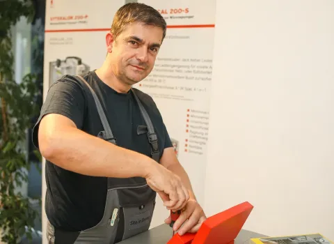 Thomas Kellenberger, ein Mann mittleren Alters mit kurzen, dunklen Haaren und leichtem Bartschatten, trägt ein schwarzes T-Shirt und eine graue Latzhose mit der Aufschrift "Sito in Broou". Er lächelt leicht in die Kamera und hält einen Schraubenzieher in der Hand, während er an einem roten Objekt auf einer grauen Oberfläche arbeitet. Im Hintergrund ist ein weiß-rotes Schild mit der Aufschrift "CAL 200-S Wärmepumpen" und grüne Pflanzen zu erkennen.