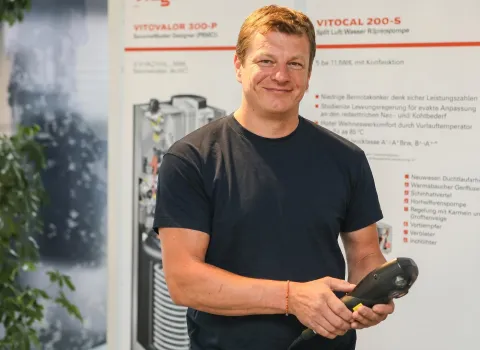 Uwe Schieck, ein lächelnder Mann mit kurzen, hellbraunen Haaren, trägt ein dunkelblaues T-Shirt und hält ein schwarzes Handgerät in seinen Händen. An seinem rechten Handgelenk ist ein orangefarbenes Band zu sehen. Im Hintergrund sind unscharfe Produktschilder von Viessmann (Vitovalor 300-P, Vitocal 200-S) und technische Illustrationen erkennbar.