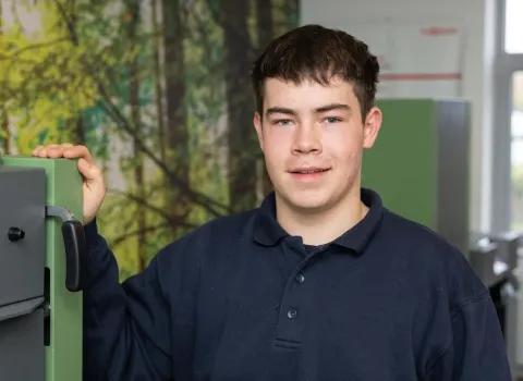 Junger, lächelnder Techniker in dunkelblauem Poloshirt, seine Hand auf einem grünen Heizsystem-Modul. Hintergrund mit Waldmotiv und Viessmann-Logo deutet auf nachhaltige Energie und Ausbildung hin.