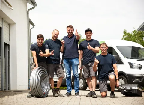 Fünf Mitarbeiter eines SHK-Betriebs stehen gut gelaunt vor der Werkstatt und den Firmenfahrzeugen. Sie tragen Arbeitskleidung, halten Rohrmaterial und Werkzeug und zeigen den Daumen nach oben.